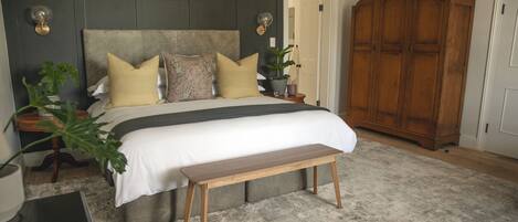 A bedroom with a large bed, a wooden bench, a wardrobe, and a nightstand with a lamp and a plant.