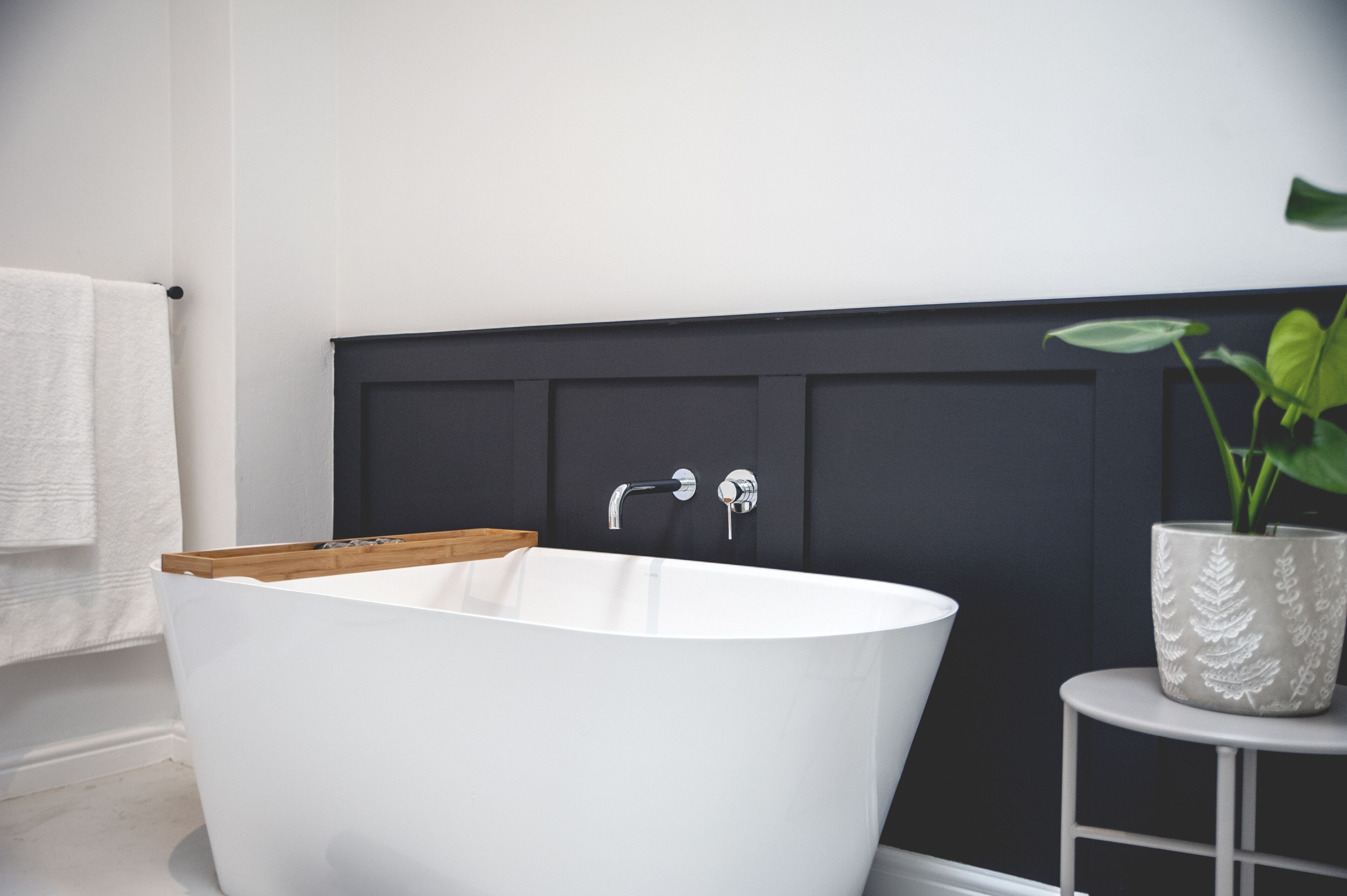 A modern bathroom with a freestanding bathtub, a wooden tray, a potted plant, and a white towel on the wall.
