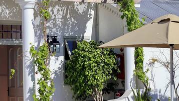 A white building with a covered outdoor seating area, a pergola with climbing plants, and a large umbrella providing shade.