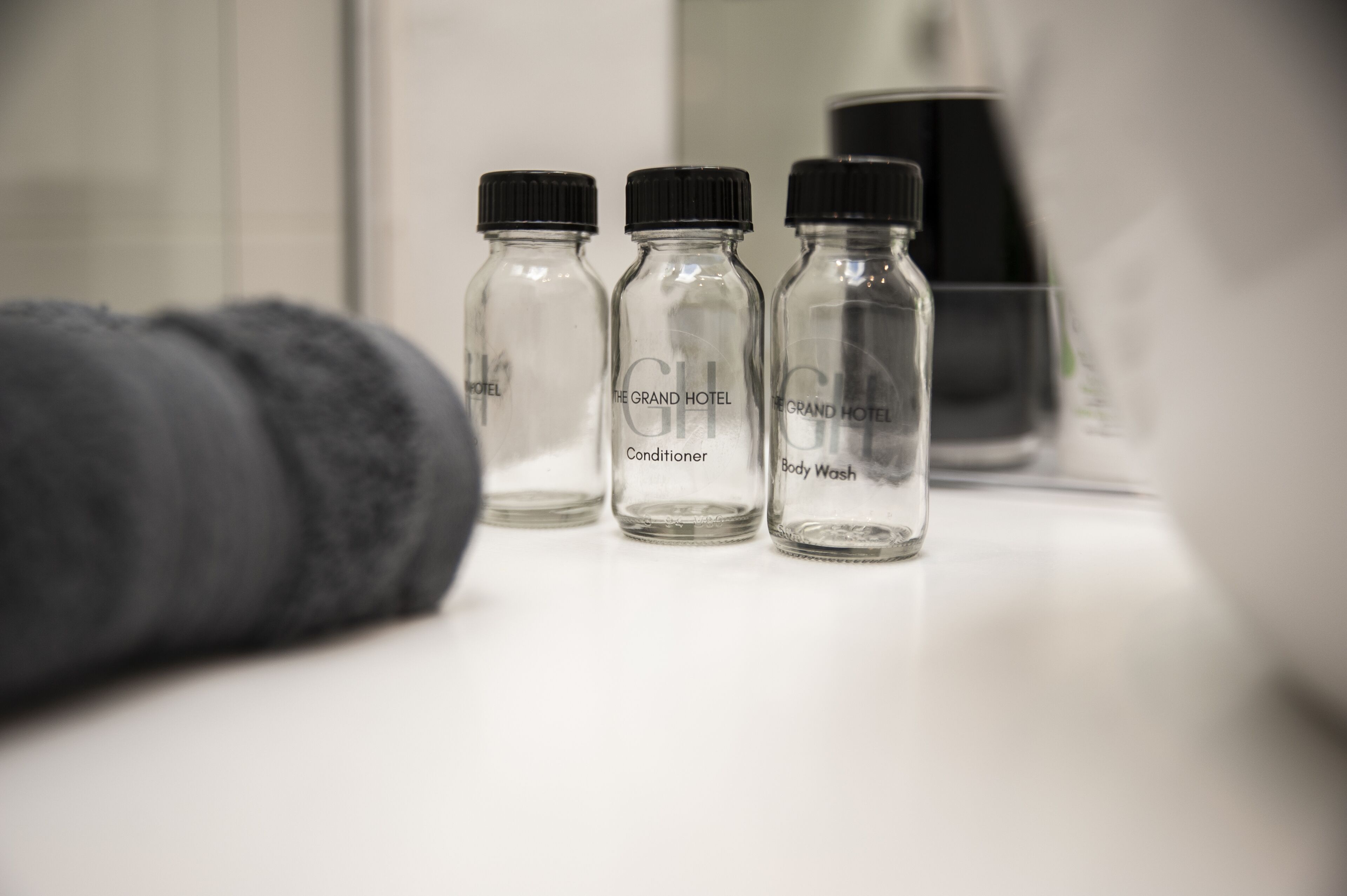Três garrafas de vidro com os dizeres 'The Grand Hotel Conditioner' e 'Body Wash' sobre uma superfície branca.