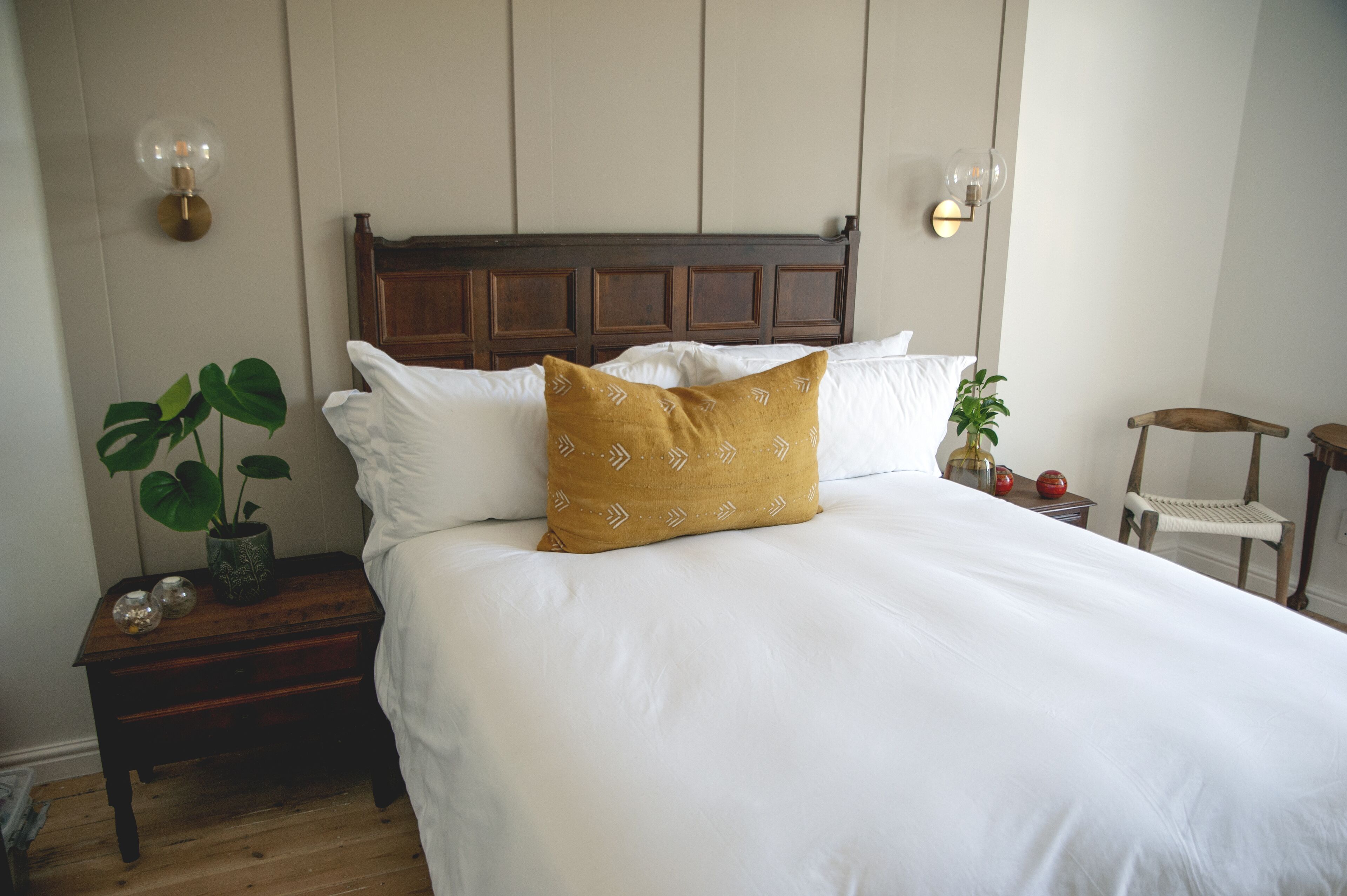 Una cama bien hecha con sábanas blancas y una sola almohada color mostaza, una mesita de noche con una planta en maceta y adornos, y una silla de madera en la esquina.