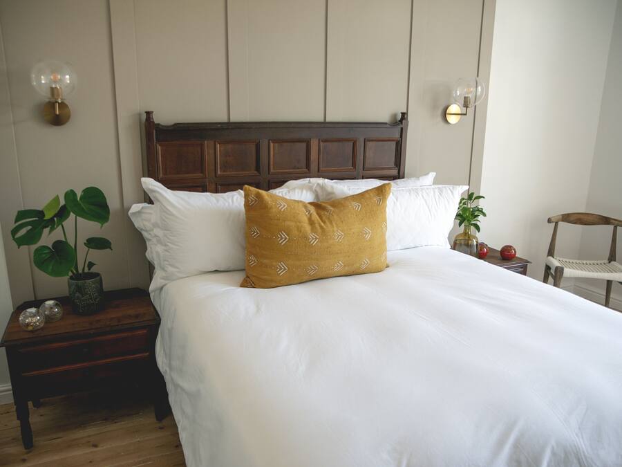 Una cama bien hecha con sábanas blancas y una sola almohada color mostaza, una mesita de noche con una planta en maceta y adornos, y una silla de madera en la esquina.