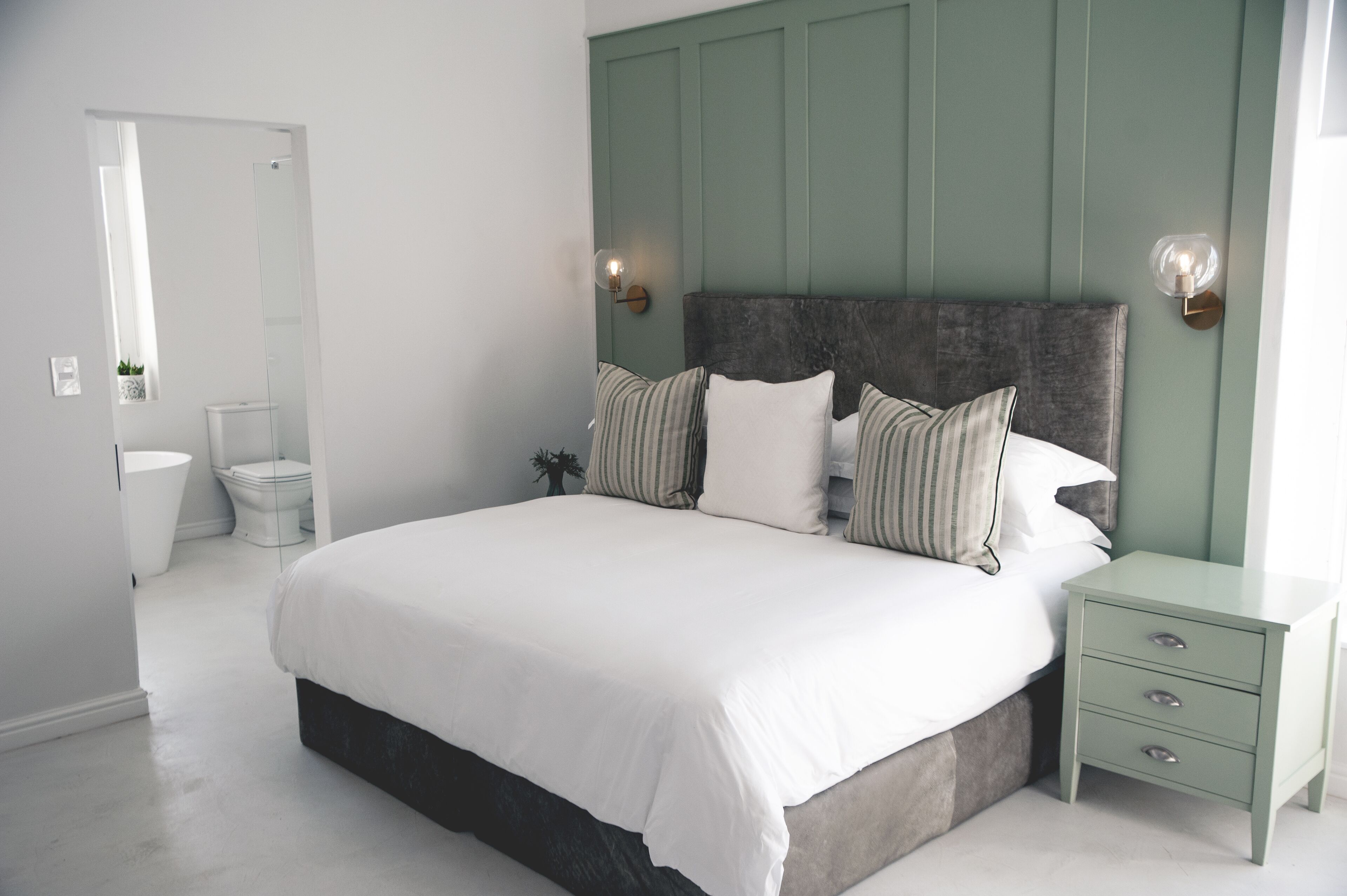 A bedroom with a large bed, a nightstand with drawers, a green headboard, and a wall-mounted light fixture.