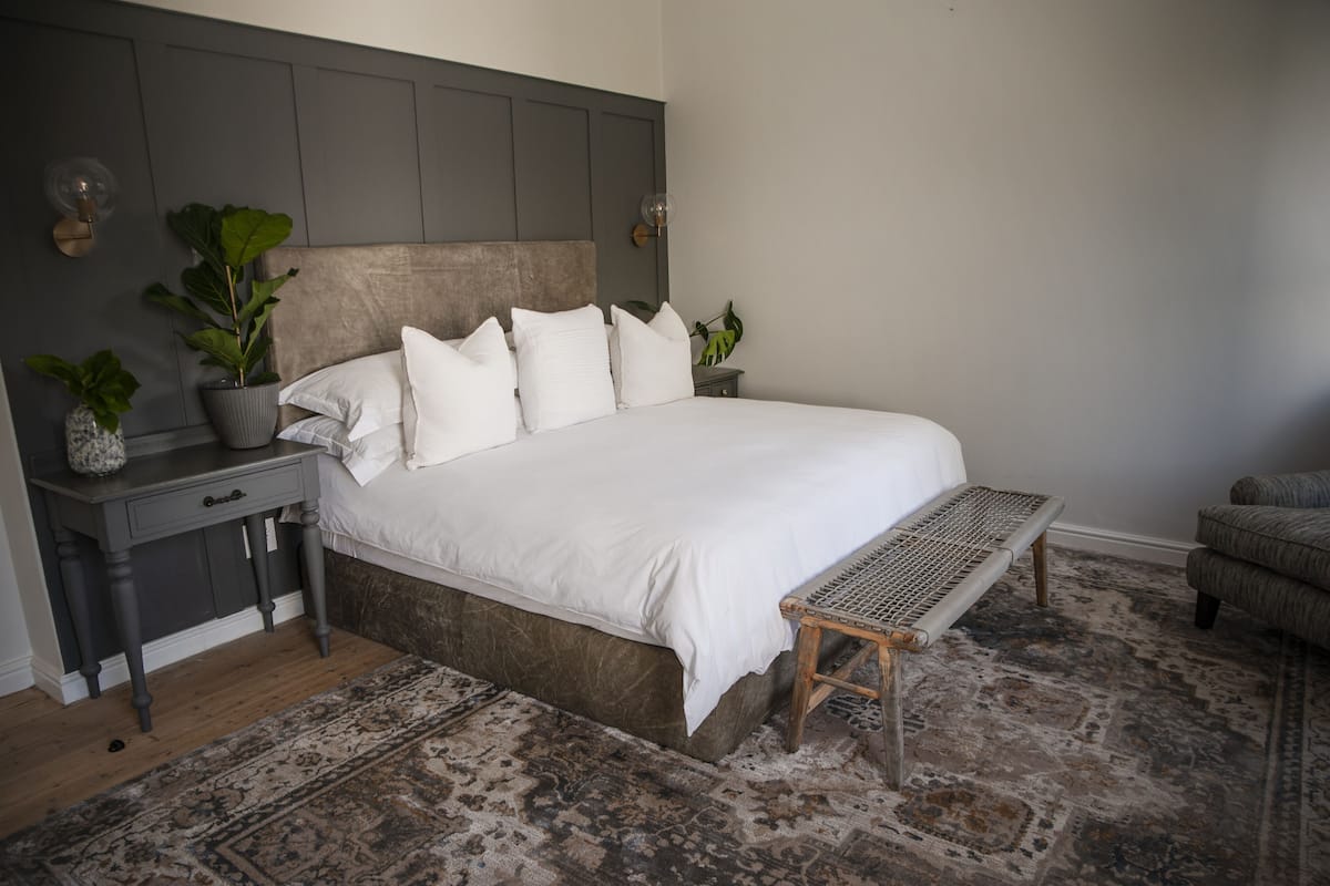 A bedroom with a large bed, a bedside table with a lamp, two potted plants, and a bench.