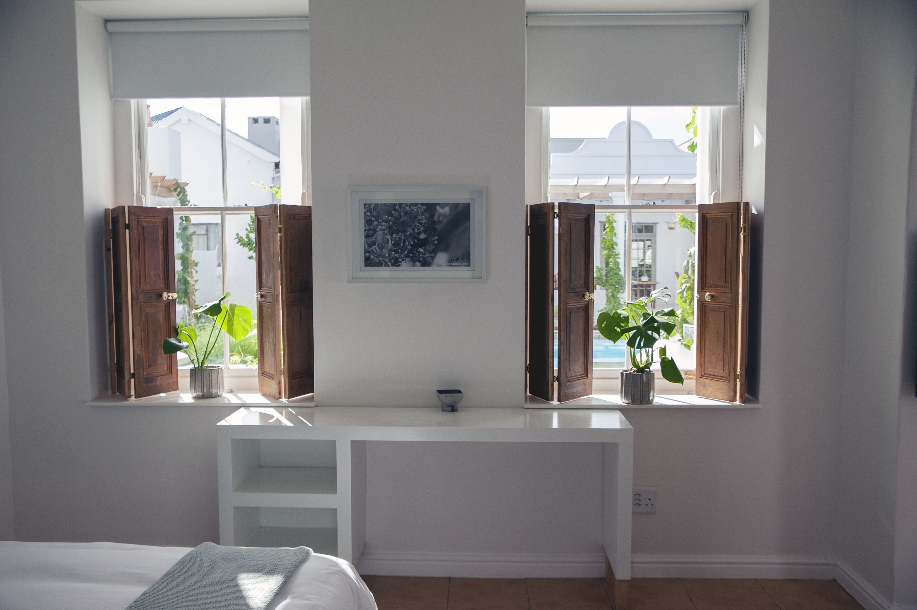 Un dormitorio con una cama blanca, un escritorio, dos ventanas con contraventanas abiertas y plantas en macetas.