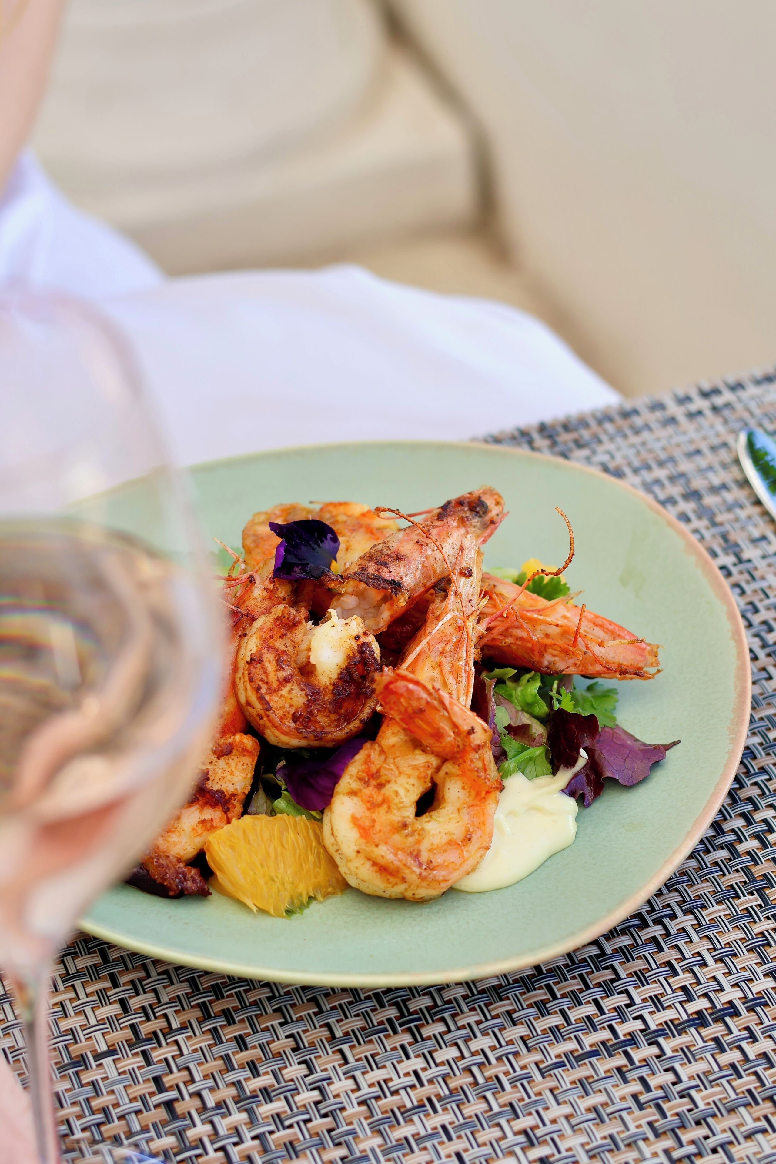 A plate of grilled shrimp with vegetables and a glass of white wine on a woven table.
