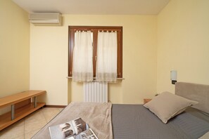 Una camera da letto con un letto, un comodino, una panca, una finestra con tende e un condizionatore d'aria.