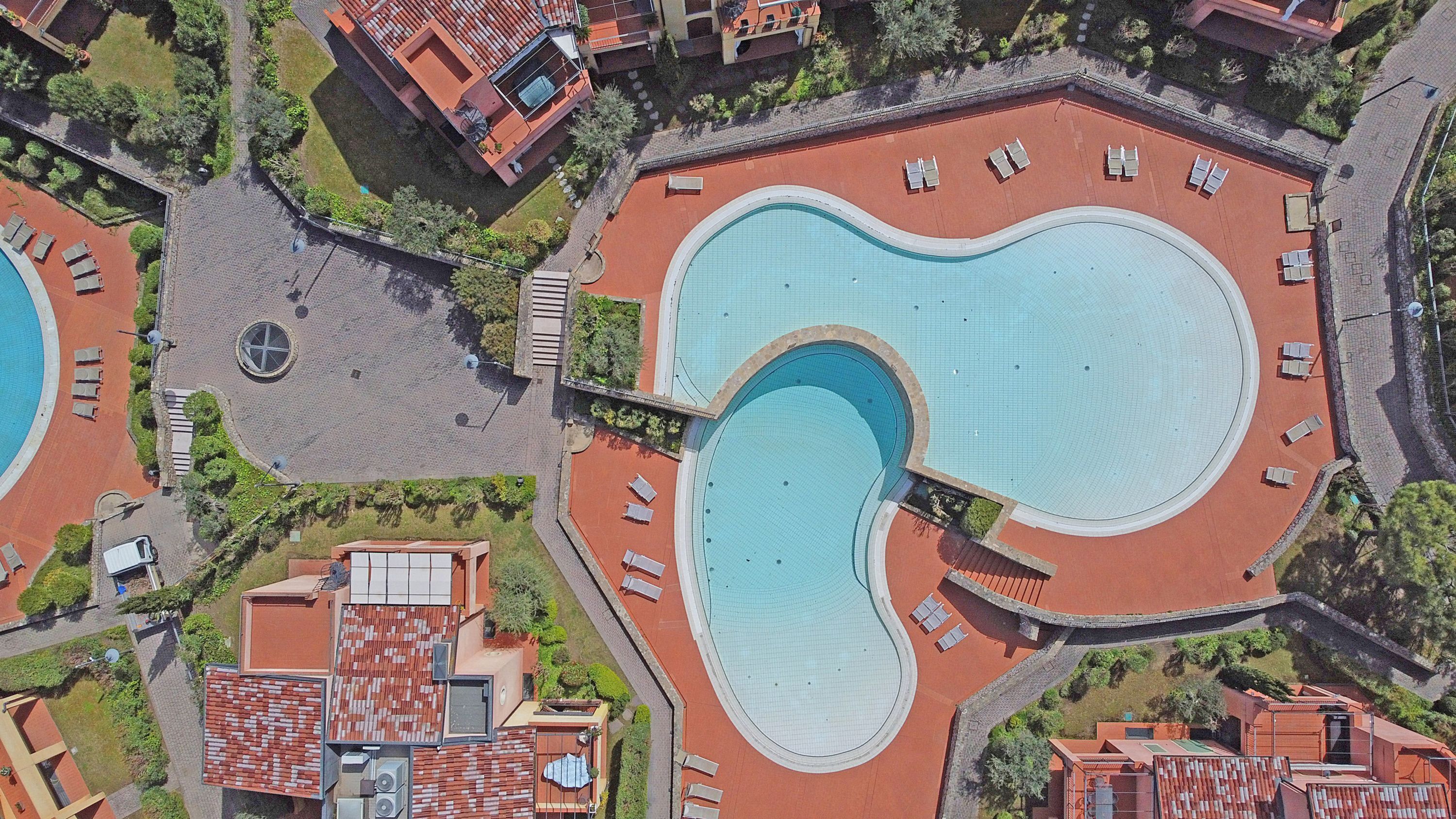 Vista aérea de un complejo turístico con dos piscinas grandes, un paseo techado y edificios circundantes.