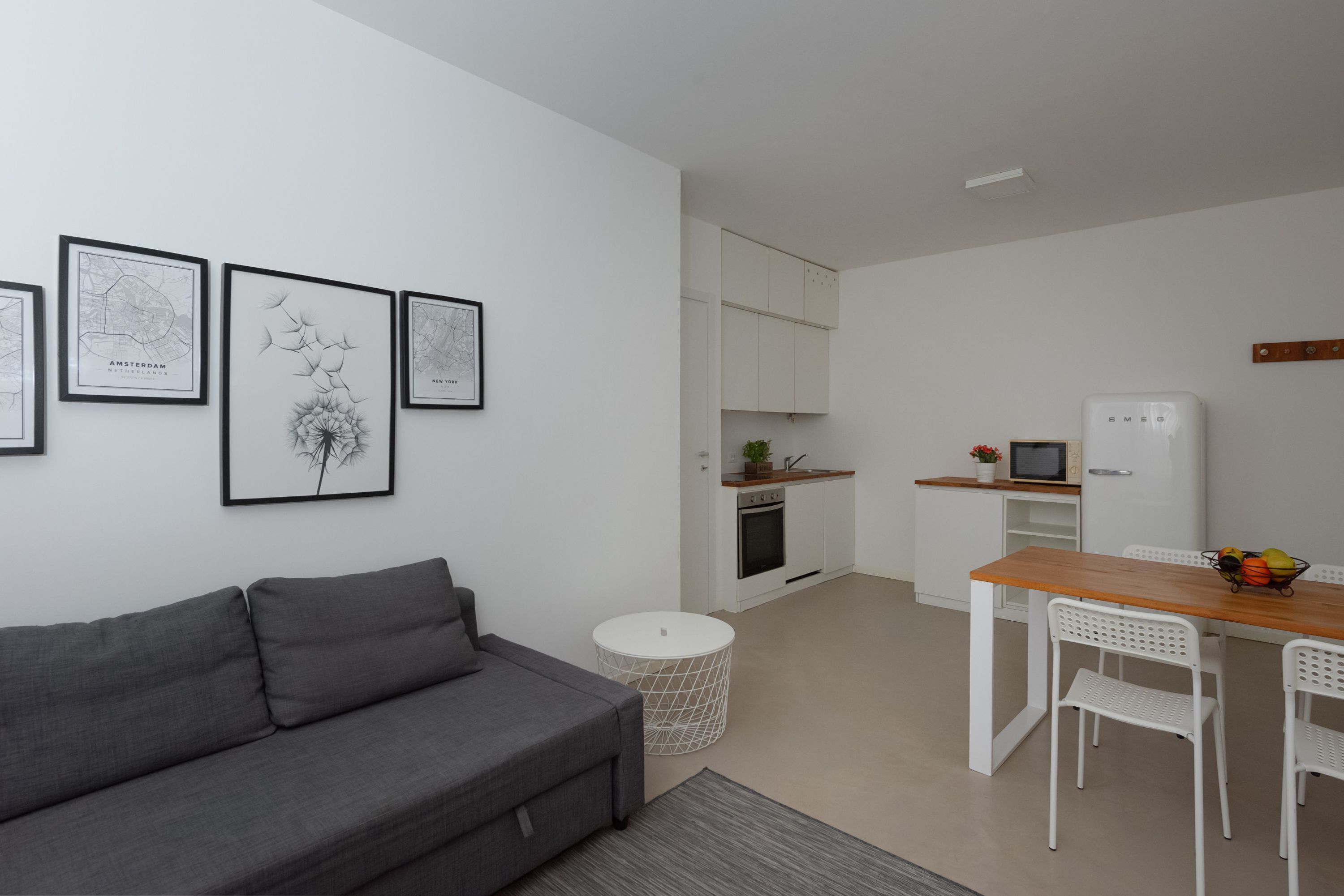 A modern living room with a grey sofa, a small round table, and a white dining table with chairs. There are framed artworks on the wall and a kitchen area with white cabinets and appliances.