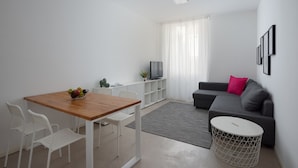 A modern living room with a wooden dining table, white chairs, a grey sofa, a TV, and a small round table. - Residence Orti 18 by Wonderful Italy (Salò)