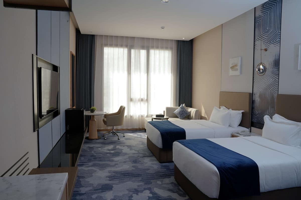 Habitación de hotel con dos camas, un escritorio, una silla, un televisor y un amplio ventanal con cortinas.