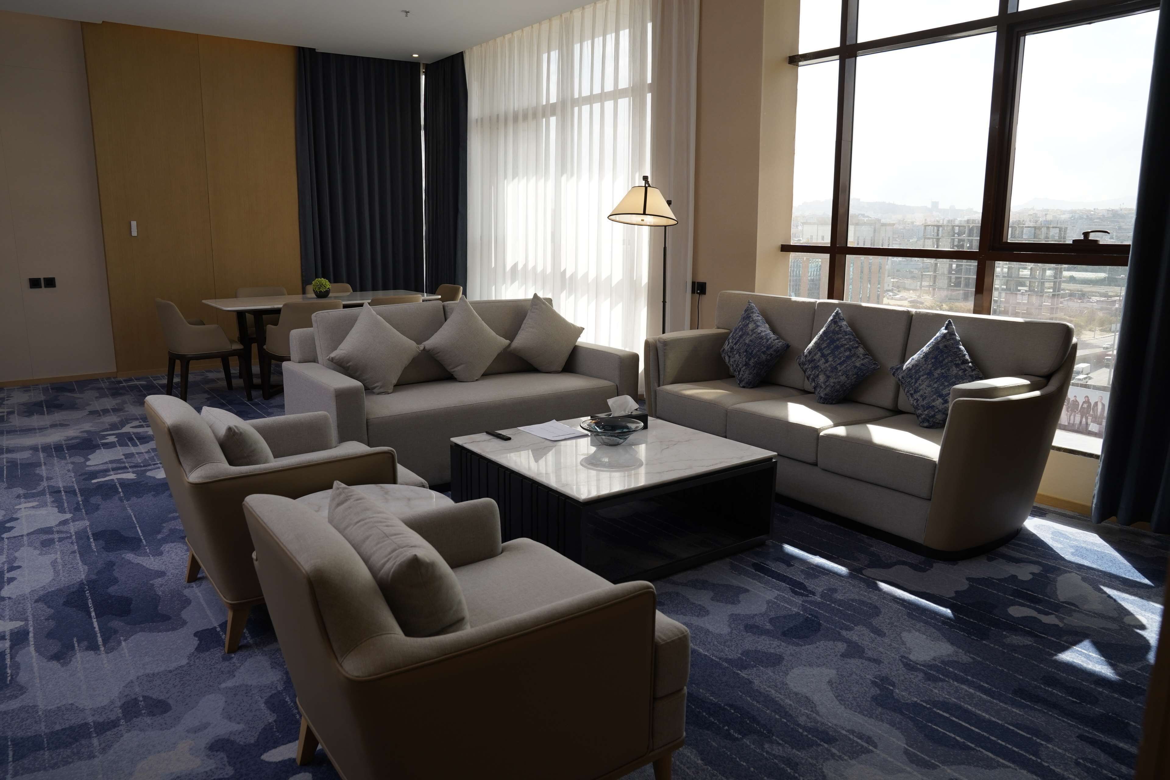 A modern hotel room with a large window, a sofa set, a coffee table, and a dining area.