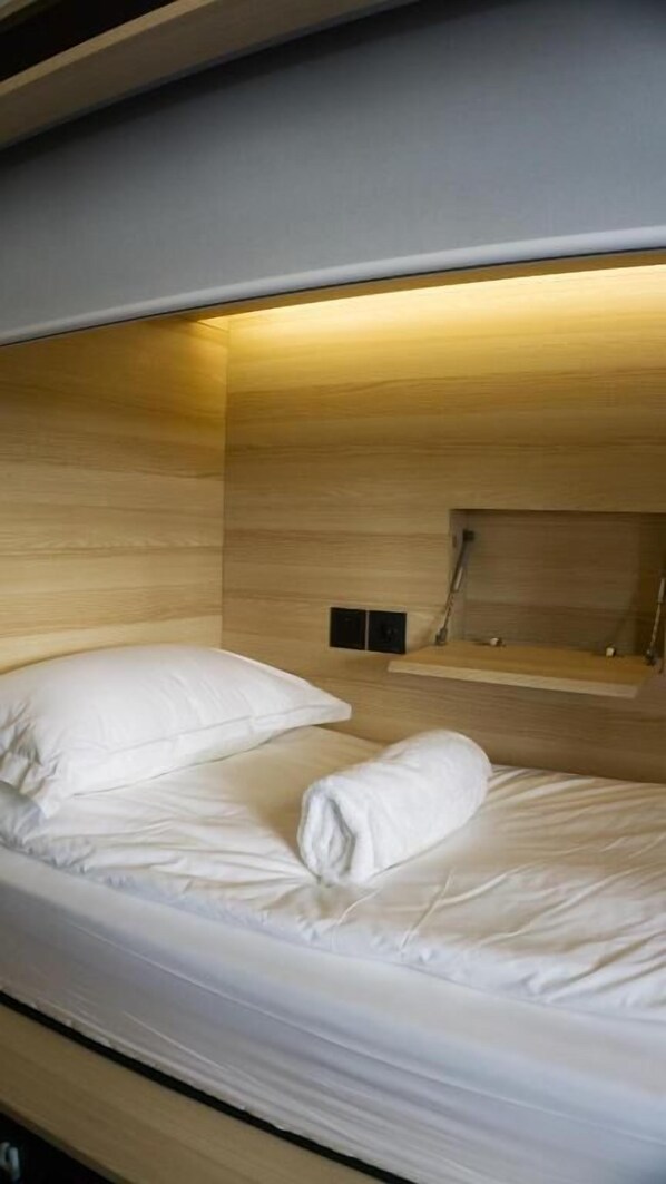 Una cama litera con colchón y almohadas blancas, un cabecero de madera y una pequeña estantería con un espejo y una toalla.
