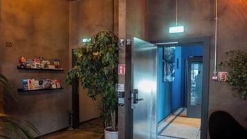 A hallway with a potted plant, a metal elevator, and a wall-mounted shelf with books.