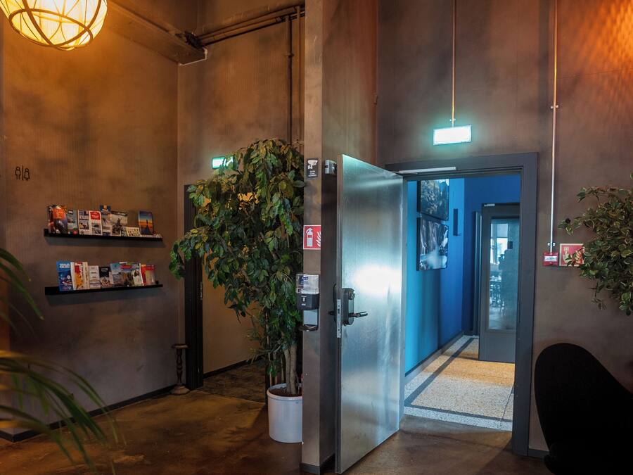 A hallway with a potted plant, a metal elevator, and a wall-mounted shelf with books.