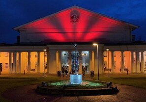 Ein Gebäude mit einer zentralen Fontäne und einer rot beleuchteten Fassade im Abenddämmerung.