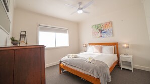 Une chambre à coucher comprenant un lit, des tables de chevet, un ventilateur de plafond, une fenêtre et un tableau.