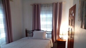 A bedroom with a bed, a nightstand, a lamp, a door, and a window with curtains. - Horse Shoe Villas A family friendly 2 bedroom villa near to it all  (Port Antonio)