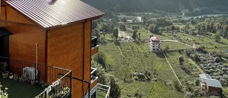 A rustic wooden building with a sloped roof, surrounded by lush greenery and a panoramic view of a valley with terraced fields and distant mountains.