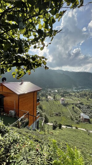 A rustic wooden building with a sloped roof, surrounded by lush greenery and a panoramic view of a valley with terraced fields and distant mountains.