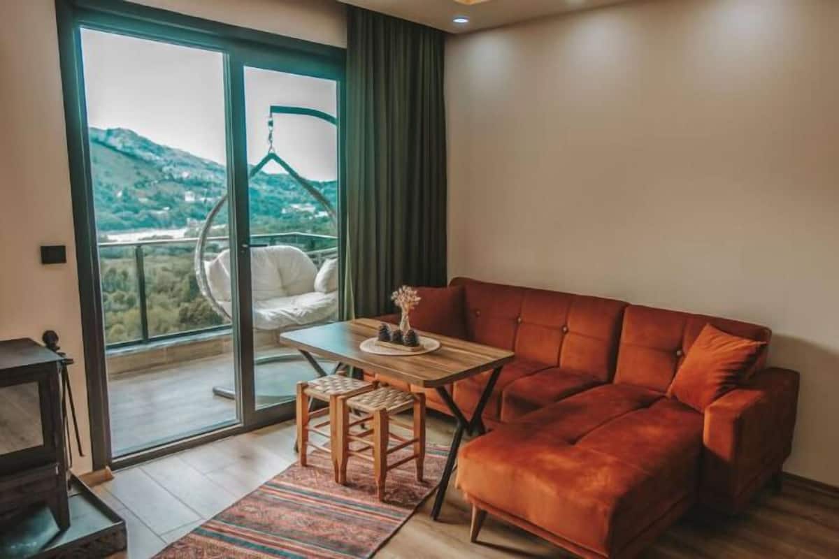 A modern living room with a sofa, a small table, and a hanging chair by a large window.