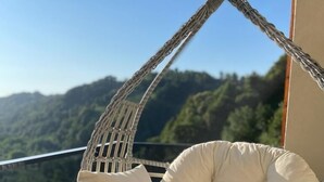 A wicker hanging chair with cushions on a balcony with a view of green hills.