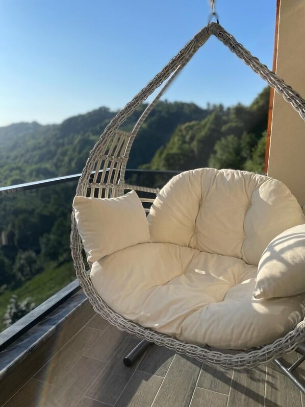 A wicker hanging chair with cushions on a balcony with a view of a forested landscape.