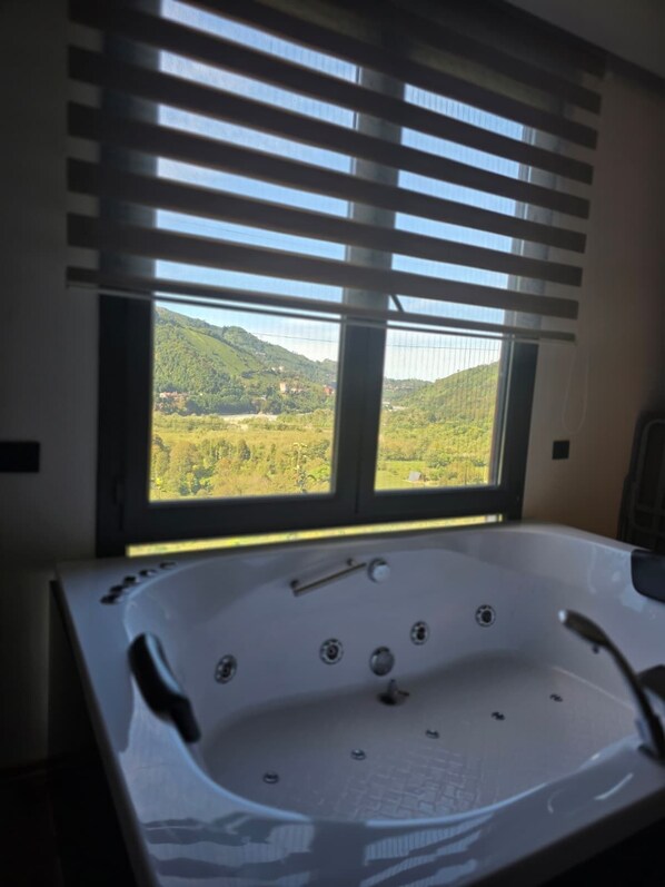 Un baño con una bañera grande, una ventana con persianas y vistas a un paisaje verde.