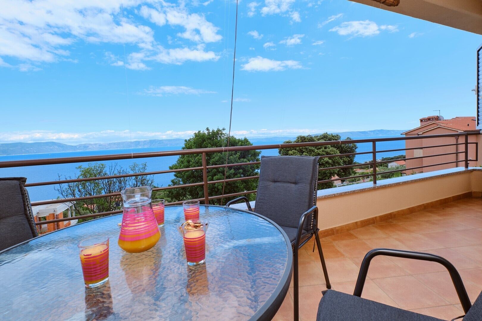 A balcony with a table, chairs, and drinks. The balcony overlooks a body of water and distant mountains.
