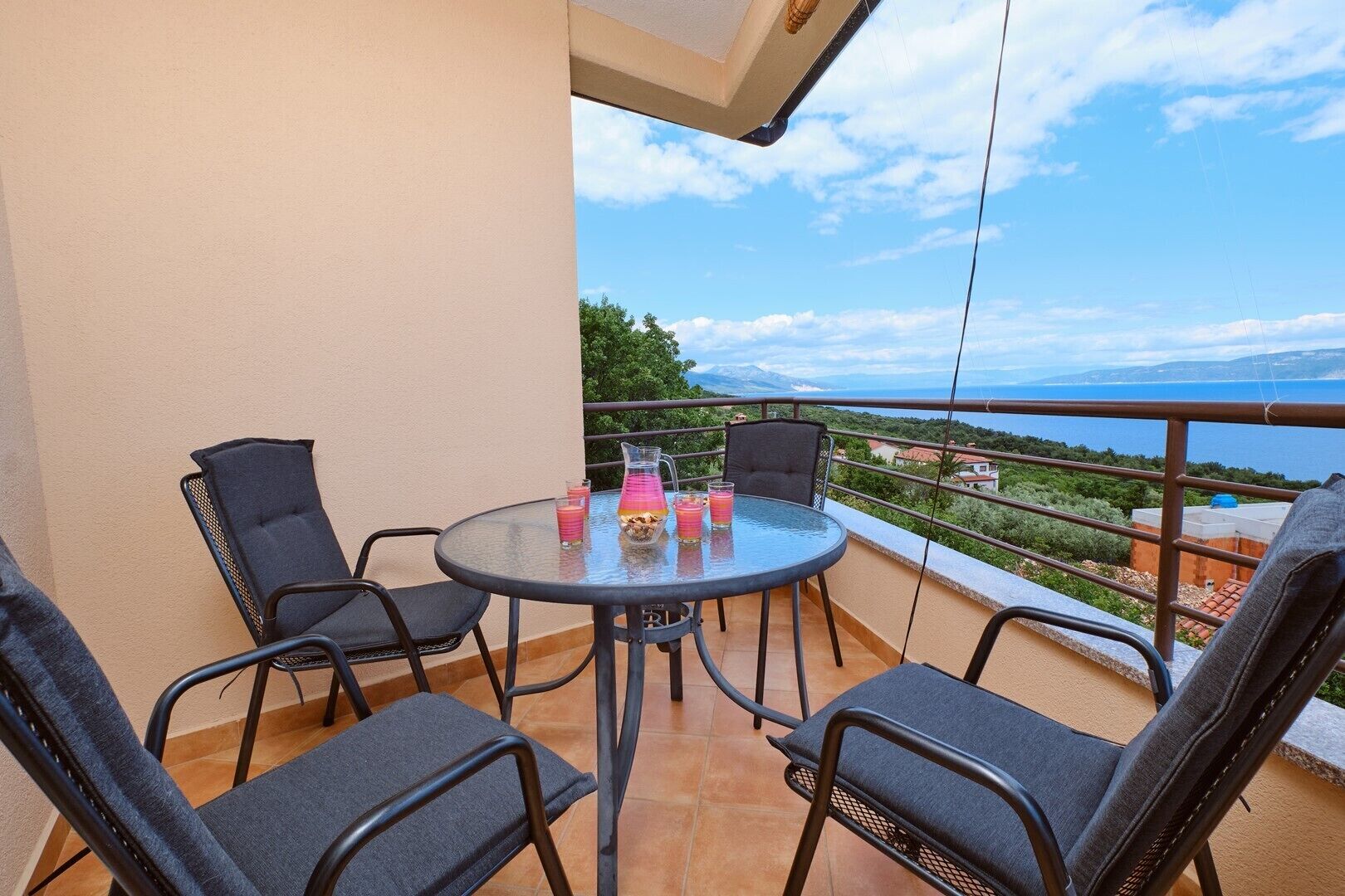 A balcony with a round table and four chairs, a pitcher of drinks, and a view of the sea and mountains.