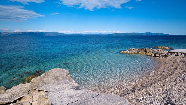 Un paisaje costero con mar azul cristalino, costa rocosa y montañas a lo lejos.