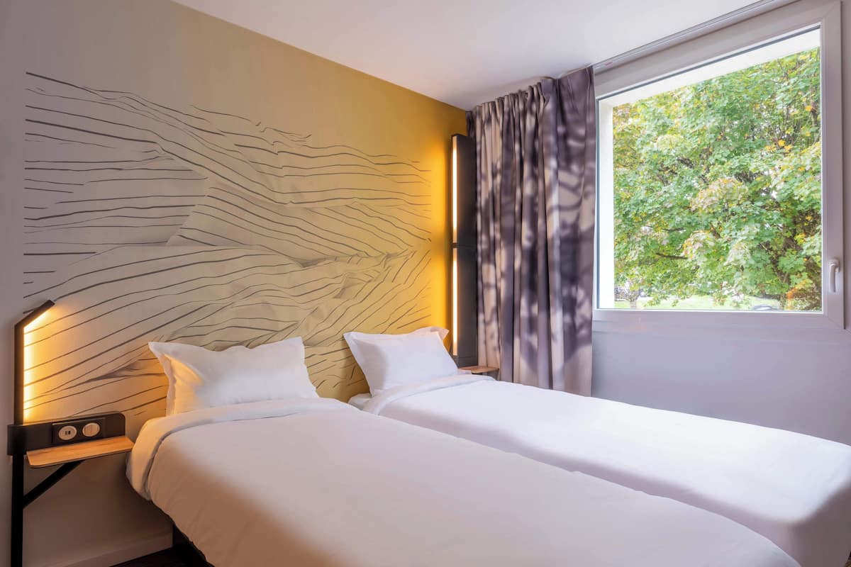 Habitación de hotel con dos camas, un cabecero moderno, una mesita de noche con lámpara y una ventana con cortinas.