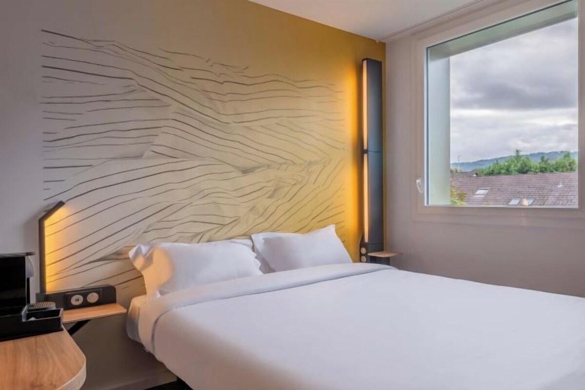 Ett modernt hotellrum med en stor säng, en sänggavel med en unik design, ett nattduksbord med en radio och ett fönster med utsikt över staden.