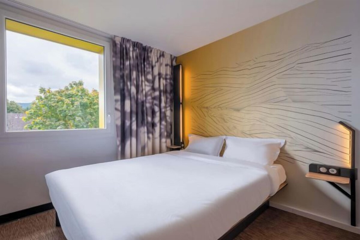 A hotel room with a large bed, a window with curtains, a wall-mounted light, and a view of trees outside.