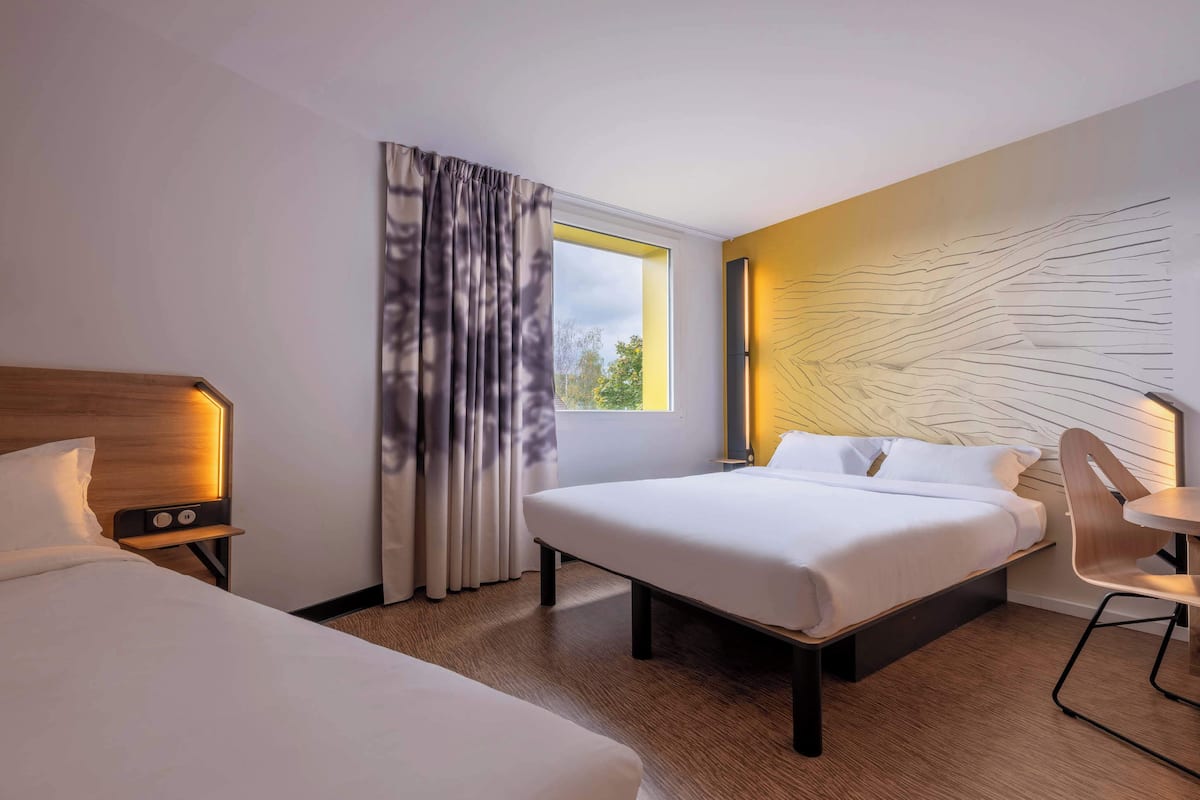 Habitación de hotel con dos camas, cabecero de madera, mesita de noche con lámpara, ventana con cortinas y pared con diseño geométrico.