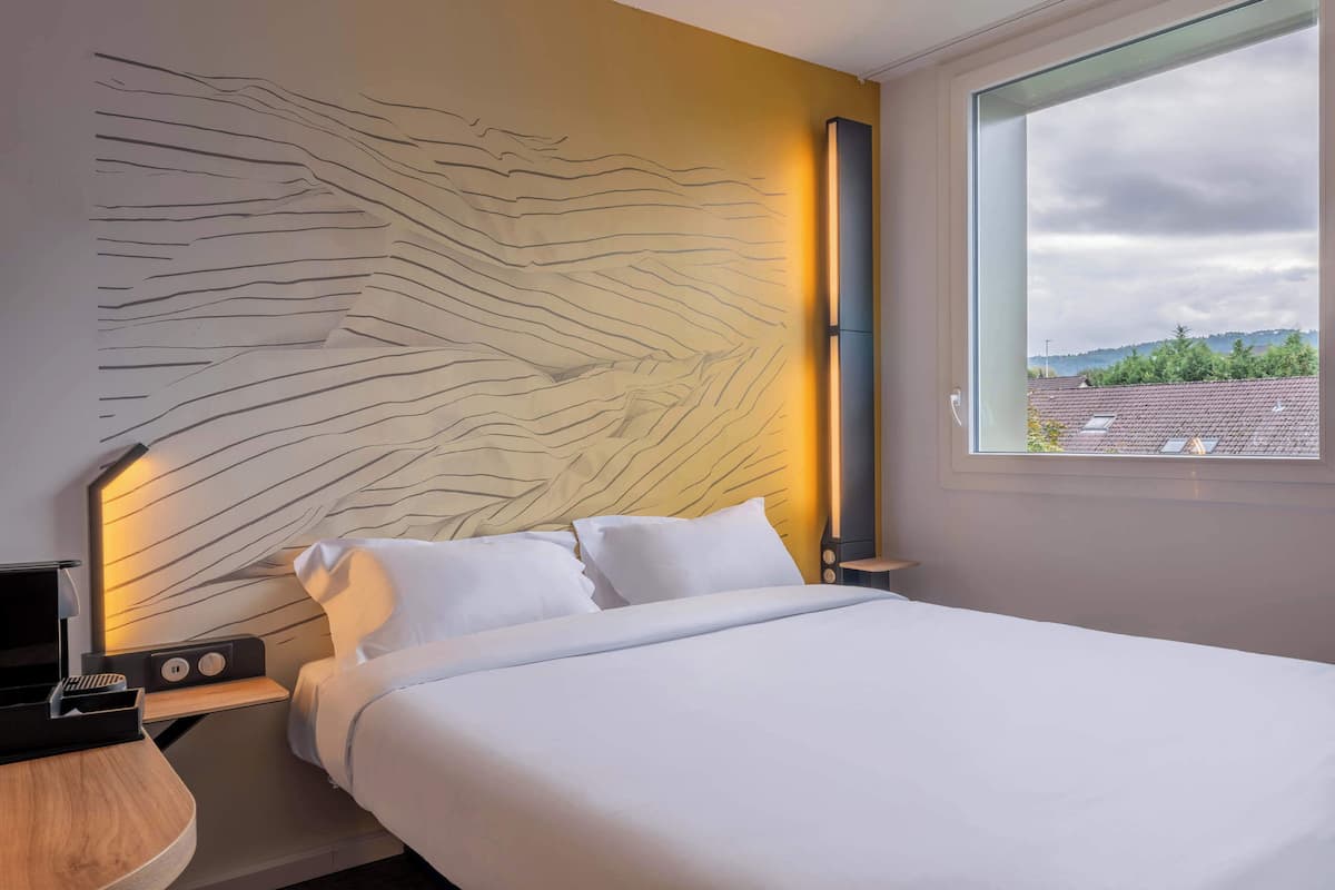 Una habitación de hotel moderna con una cama grande, una mesita de noche con lámpara y vistas a la ciudad a través de un ventanal amplio.