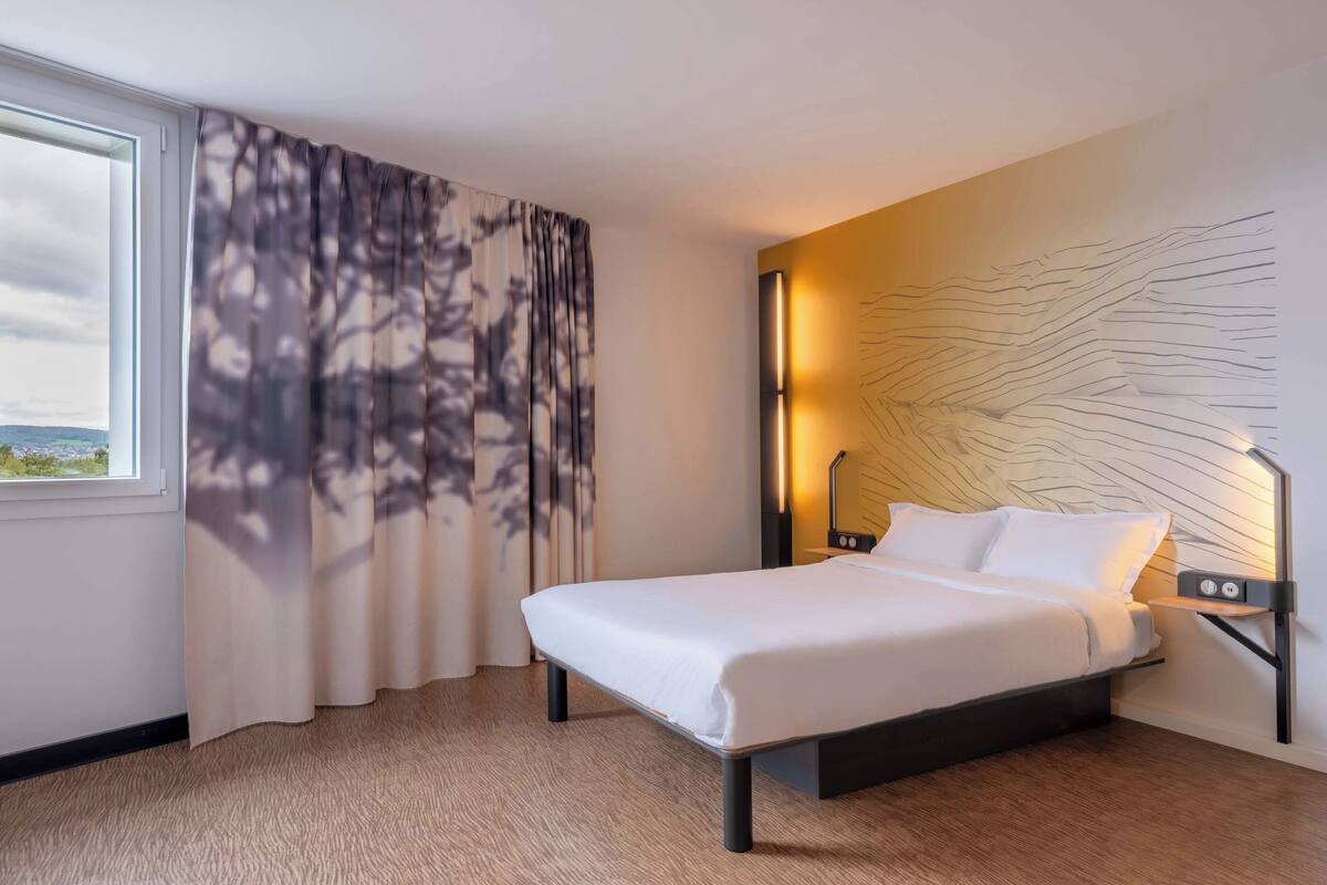 Una habitación de hotel moderna con una cama, vistas por la ventana y una lámpara fijada a la pared.