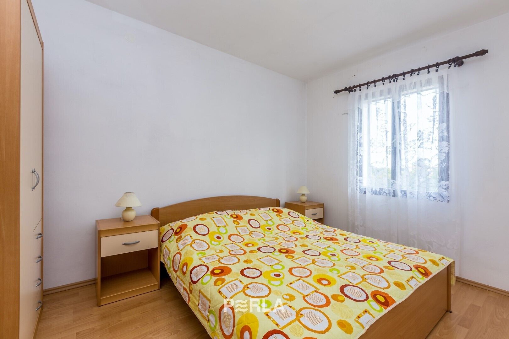 Une chambre à coucher avec un lit en bois, deux tables de chevet avec des lampes, une fenêtre avec des rideaux légers et une porte avec une poignée.