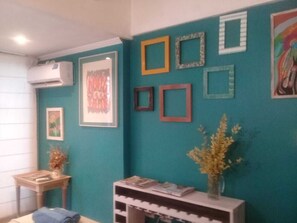 A room with teal walls, framed artwork, a small table with a vase, and a shelf with books.