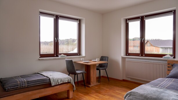 A small, tidy room with a wooden bed, a small table with chairs, and two windows offering views of the countryside.