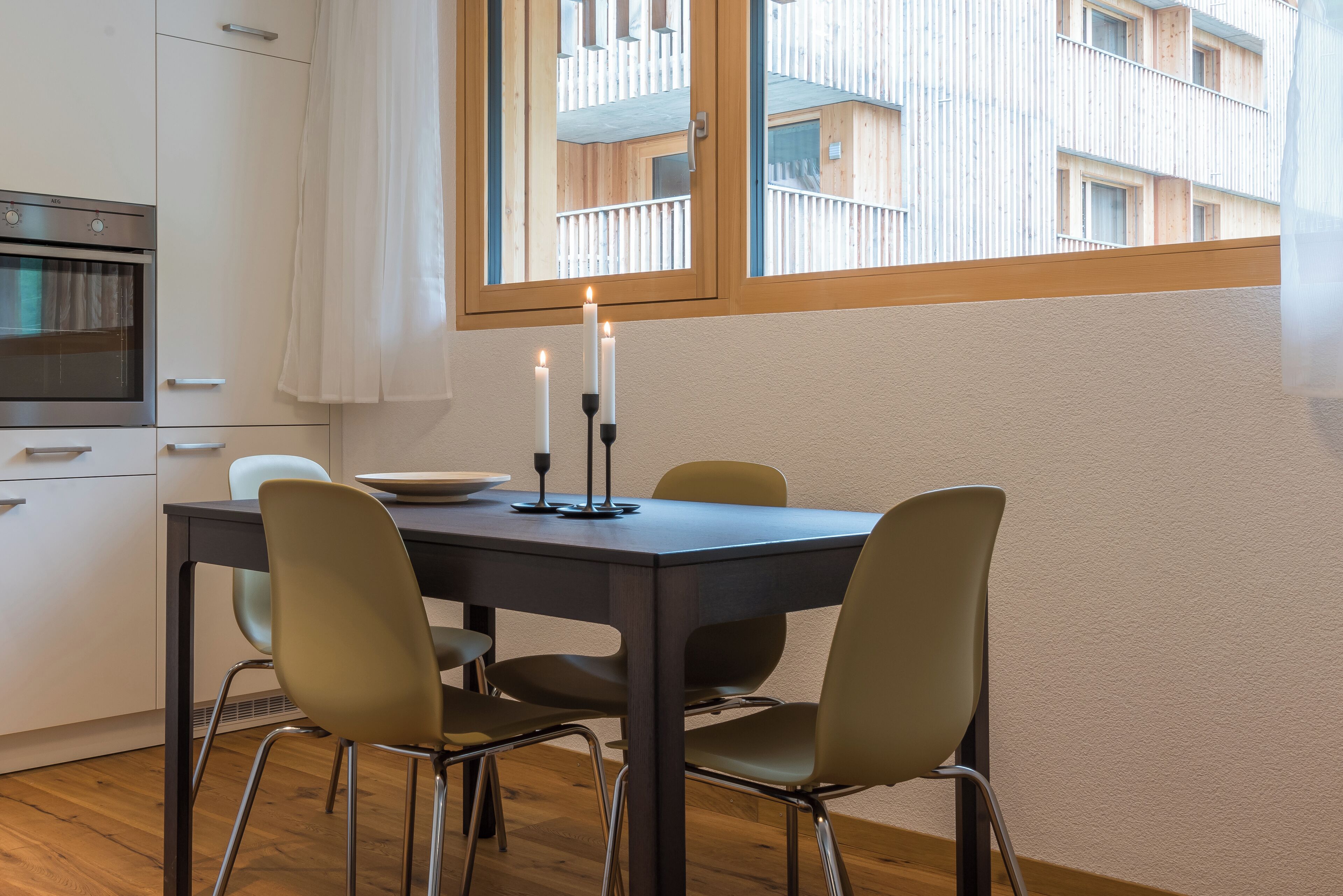 Une cuisine moderne avec une table à manger et des chaises, une fenêtre donnant sur un immeuble et une bougie sur la table.