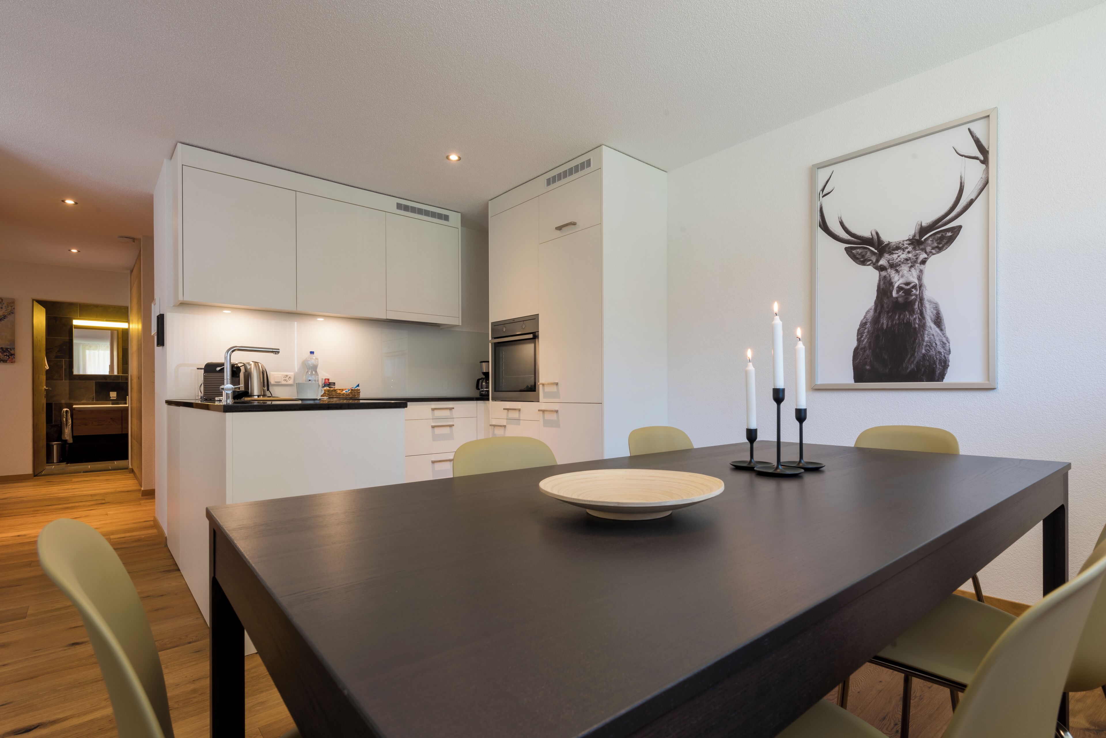 A modern kitchen with white cabinets, a dining table with a white plate and candles, and a framed deer picture on the wall.