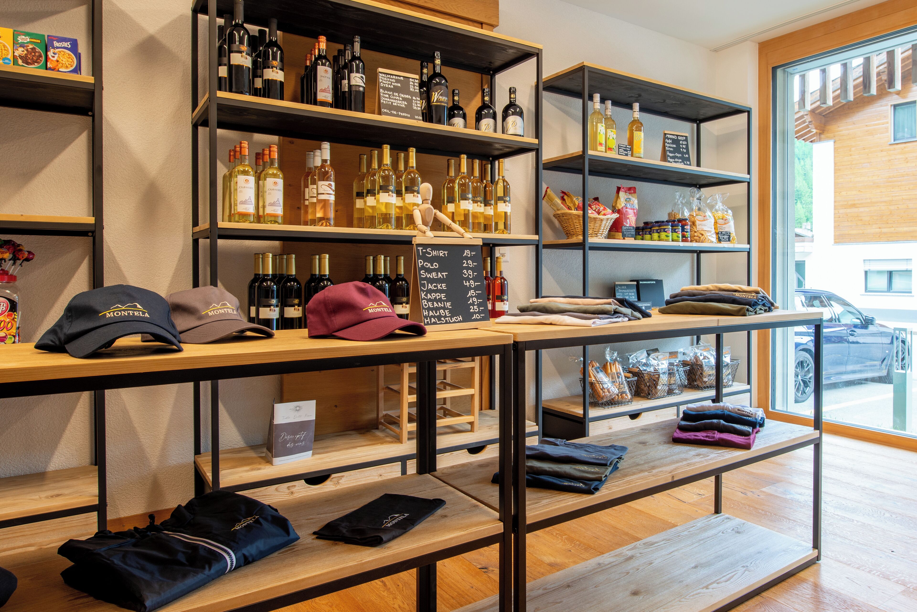A store with shelves of wine bottles, a table with folded clothes, and a window with a view of a building.