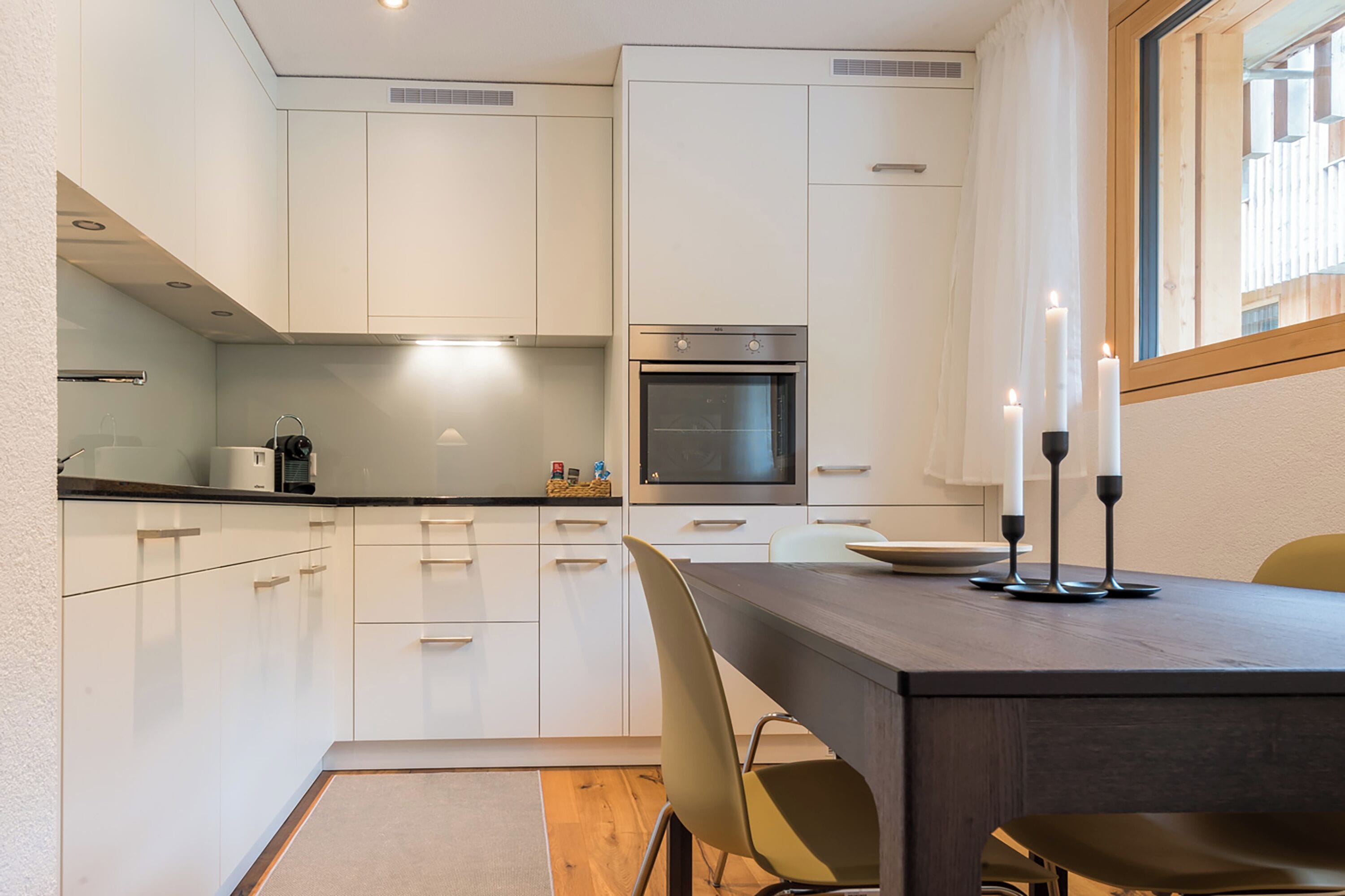 Une cuisine moderne avec des meubles blancs, un plan de travail foncé et un coin repas comprenant une table et des chaises.