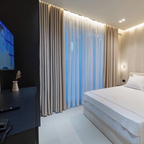 Habitación de hotel moderna con una cama grande, un televisor sobre un escritorio y una ventana con cortinas translúcidas.