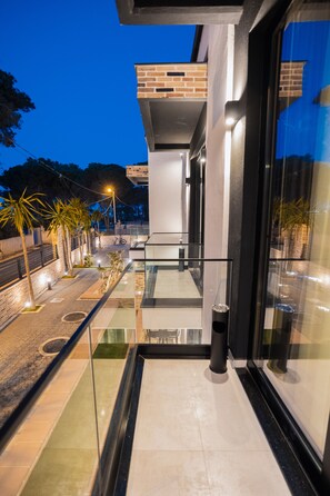 Un edificio moderno con balcón de vidrio, sección de ladrillo y un paseo empedrado bordeado de palmeras.