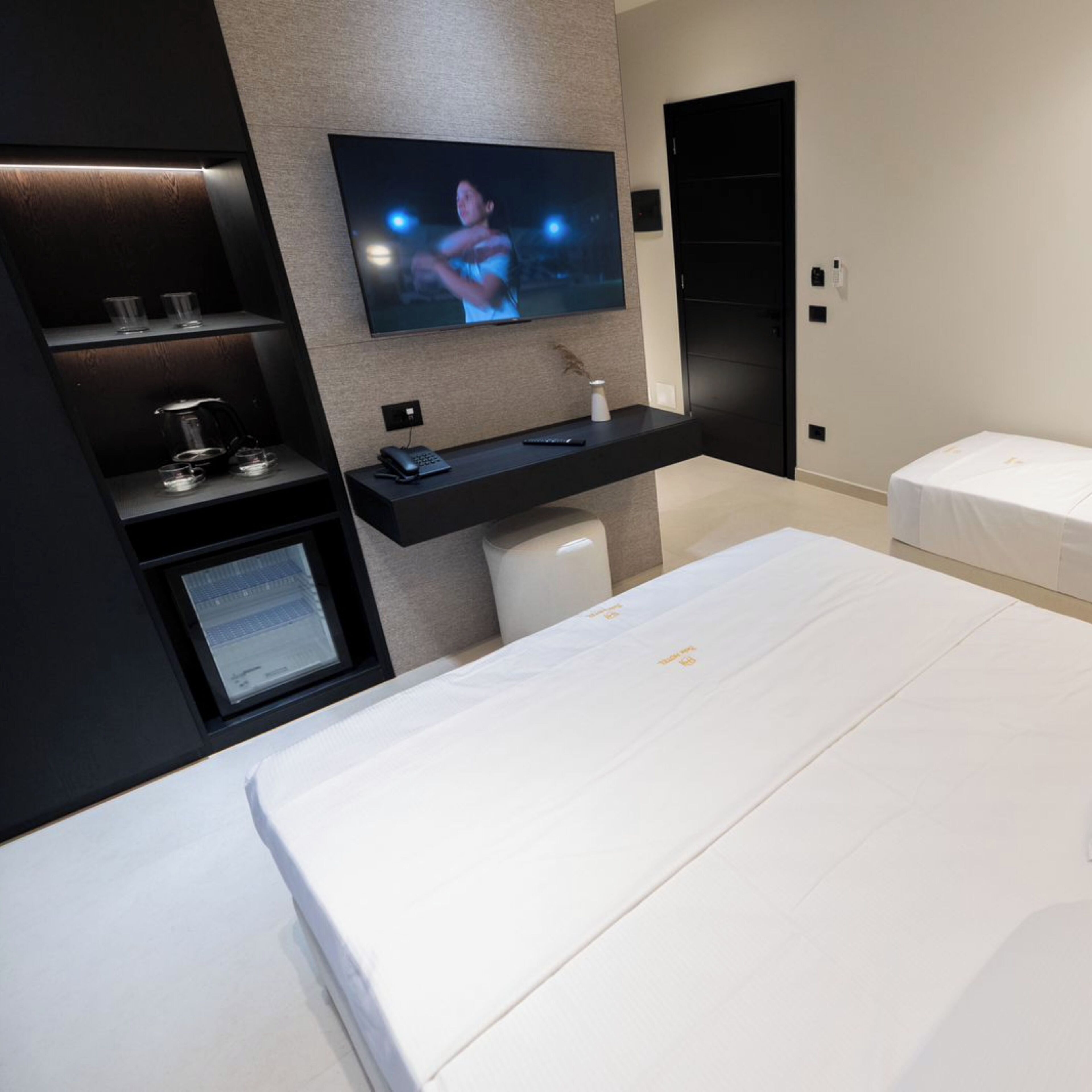 A modern hotel room with a large bed, a TV mounted on the wall, a small desk with a computer, and a bedside table with a lamp.