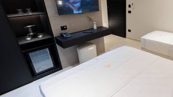 A modern hotel room with a large bed, a TV mounted on the wall, a small desk with a computer, and a bedside table with a lamp.