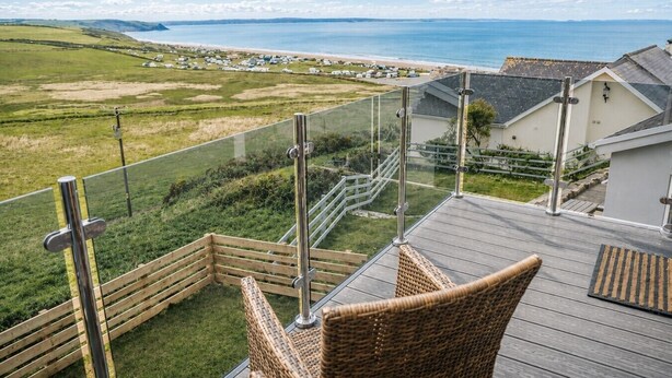 A balcony with wicker furniture, a glass railing, and a view of the sea and distant buildings. - Modern balcony property, Sea views over Newgale (Newgale)