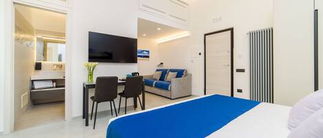 A modern hotel room with a large bed, a sofa, a TV, and a dining area with chairs and a table.